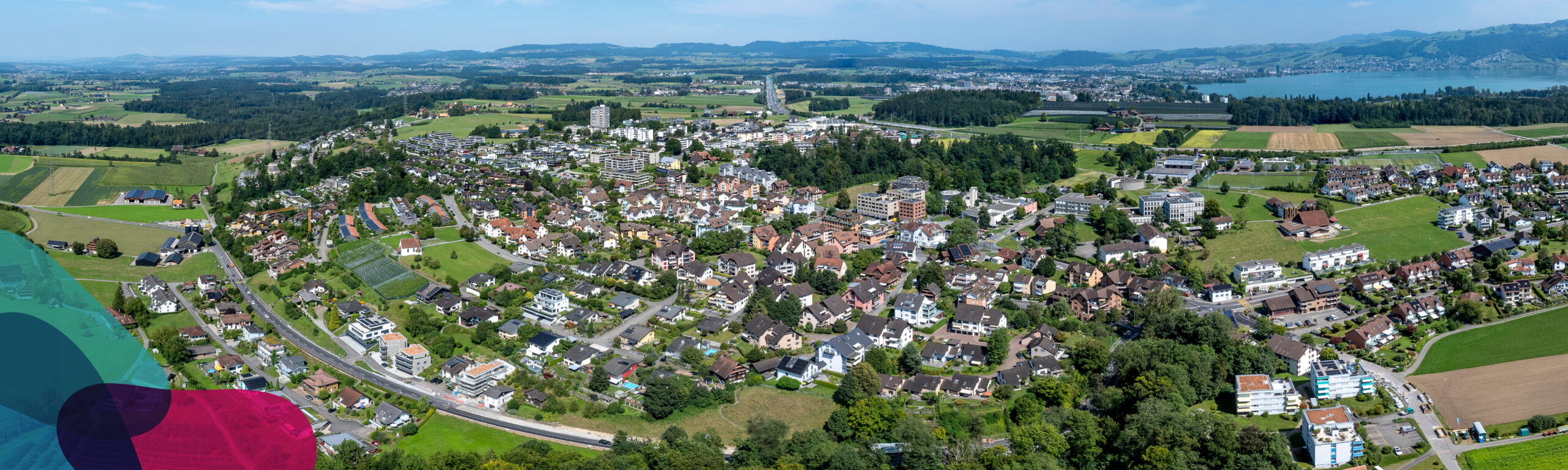Luftaufnahme der Gemeinde Hünenberg. In einer Ecke überlagern drei farbige Flächen das Bild