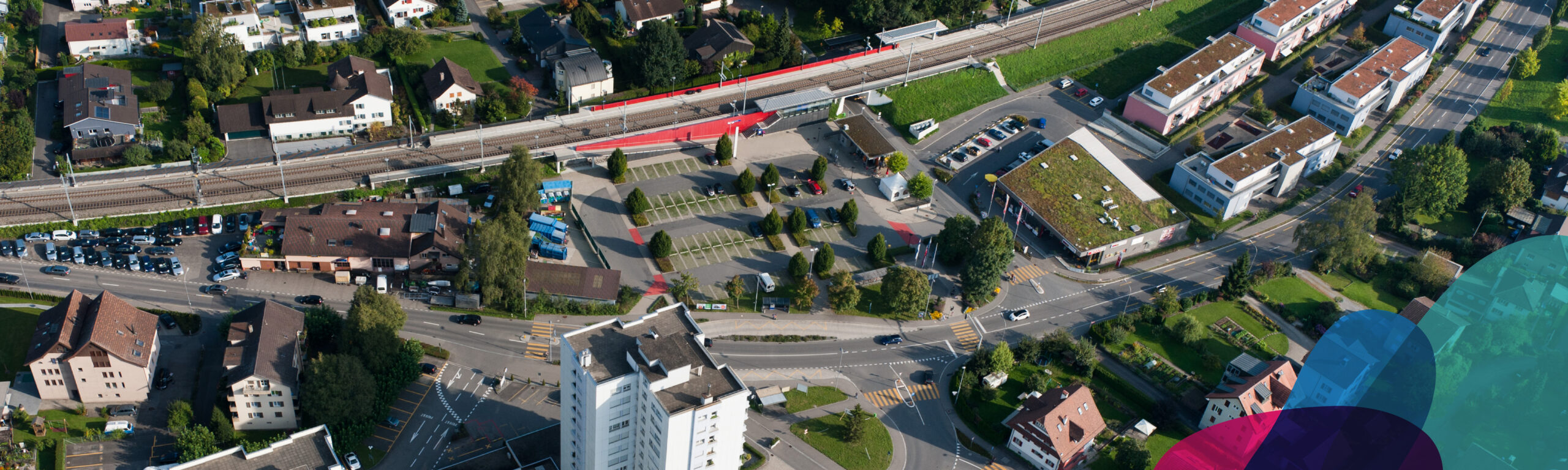 Luftaufnahme der Gemeinde Hünenberg. In einer Ecke überlagern drei farbige Flächen das Bild